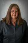 A woman with shoulder-length brown hair wearing a black shirt and a necklace with a purple pendant. She faces the camera against a plain grey background, with a serious expression.
