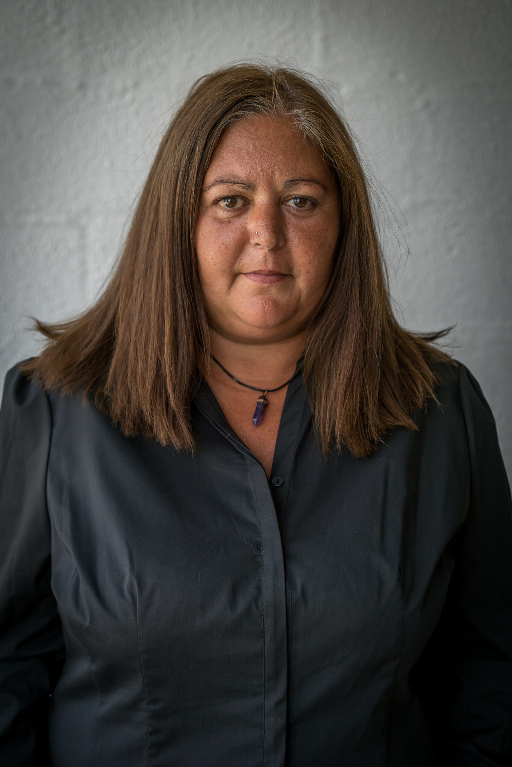 A woman with shoulder-length brown hair wearing a black shirt and a necklace with a purple pendant. She faces the camera against a plain grey background, with a serious expression.