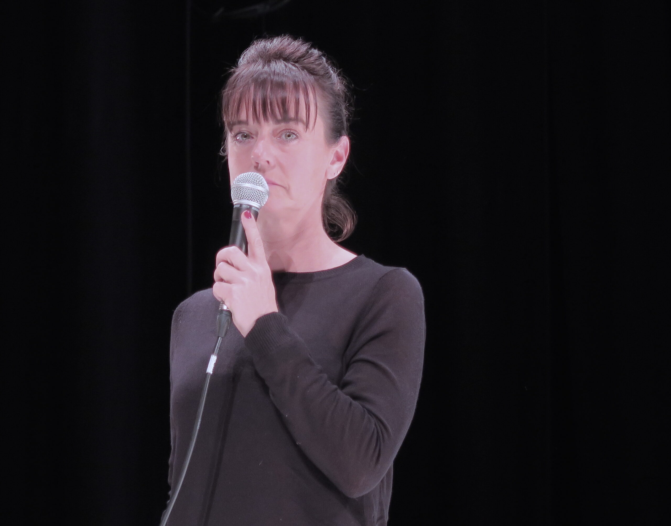 A woman in a black sweater stands on stage holding a microphone against a dark background. Her expression is focused and serious, suggesting a speech or performance.