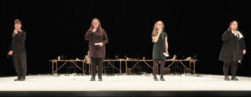 Four performers on stage, each holding microphones and wearing black, stand evenly spaced. A table with scattered objects is behind them. The scene is dramatic and focused.