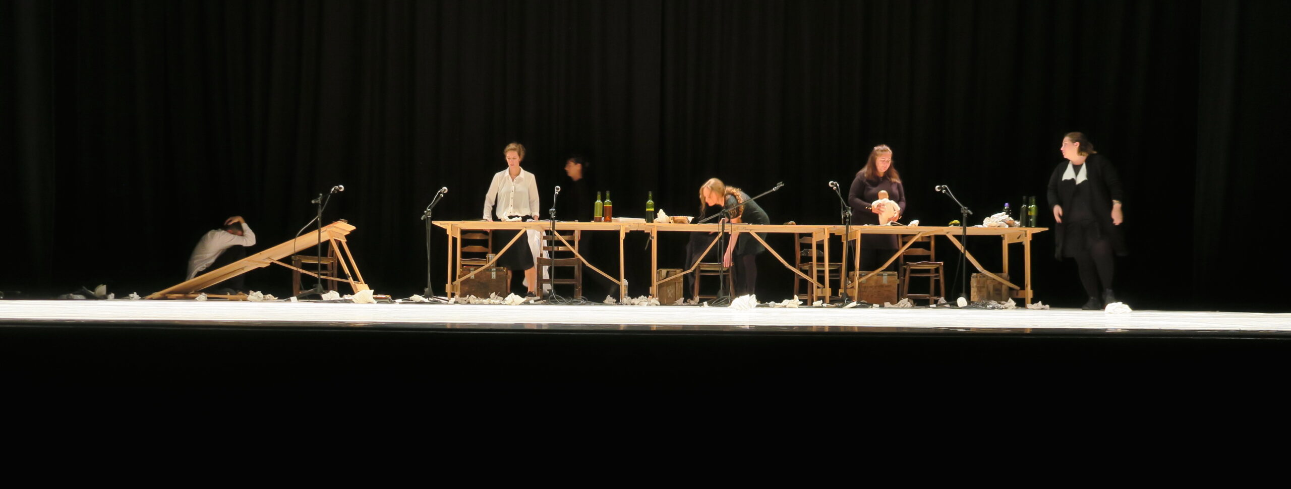 A dimly lit stage features six people in varied poses at a long table with papers and bottles. The scene conveys a dramatic and tense atmosphere.