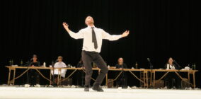 A man in a white shirt and black tie stands center stage with outstretched arms, evoking emotions. Behind him, five people sit at desks on a dimly lit stage.