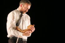 A man in a white shirt and tie cradles a baby doll tenderly against a black background, conveying a sense of care and introspection.