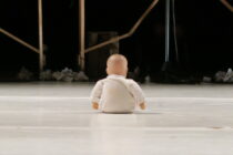 A bald doll dressed in white sits on a stage facing scaffolding, surrounded by scattered paper. The scene conveys solitude and contemplation.