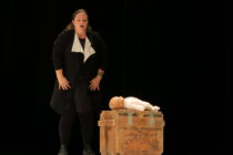 A woman on stage, wearing a black and white outfit, stands beside a wooden crate holding a baby doll. Her expression appears intense and serious.