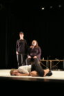 Three performers in black clothing are on stage; one stands, one sits with a microphone, and one lies curled up on the floor, conveying drama and emotion.