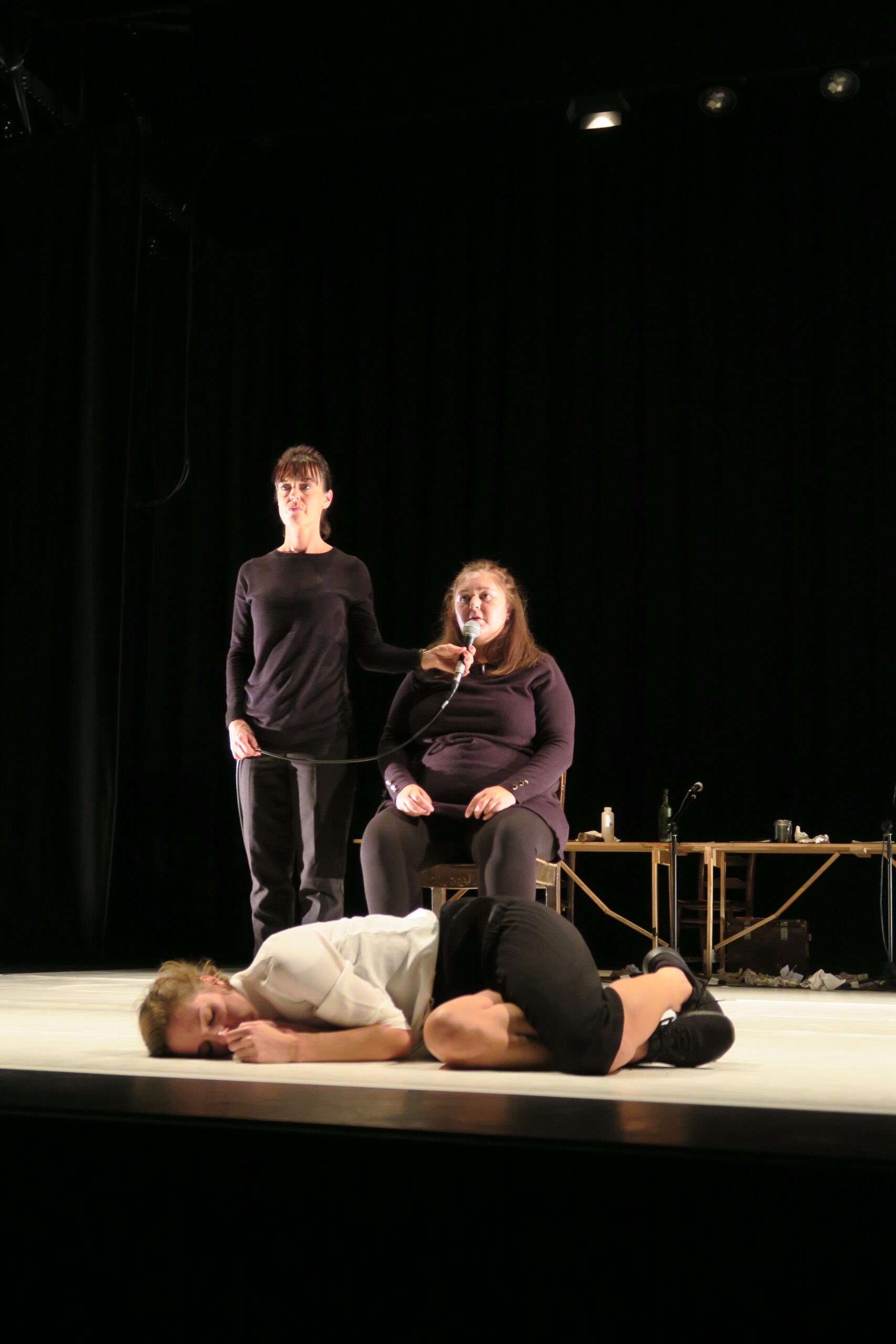 Three performers in black clothing are on stage; one stands, one sits with a microphone, and one lies curled up on the floor, conveying drama and emotion.