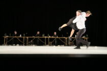 A dancer carries another in an embrace on a dimly lit stage, with four seated performers at a long table in the background, creating a dramatic scene.