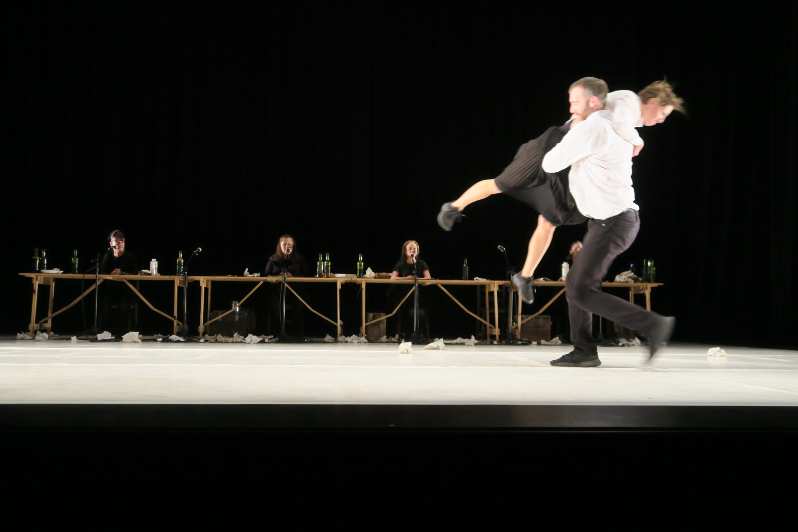 A dancer carries another in an embrace on a dimly lit stage, with four seated performers at a long table in the background, creating a dramatic scene.