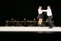 A couple dances energetically onstage, wearing formal black and white attire. In the background, six people sit at a long table, creating a lively theatrical scene.
