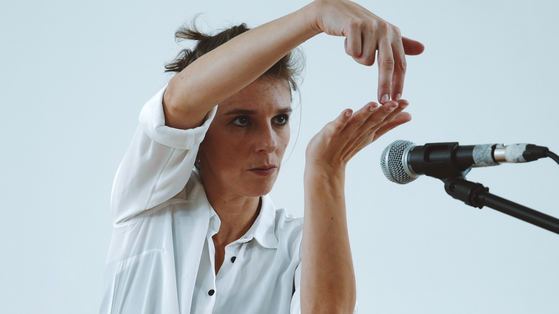 A woman in a white shirt gestures expressively with her hands near a microphone, focusing intently, set against a plain light blue background.