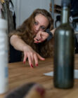 A woman with long hair is leaning across a wooden table, reaching out with one hand. Her expression is focused. A bottle and a microphone are on the table.