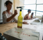 A person speaks into a microphone at a wooden table with a first aid case and bottle in focus. The setting is relaxed and casual.
