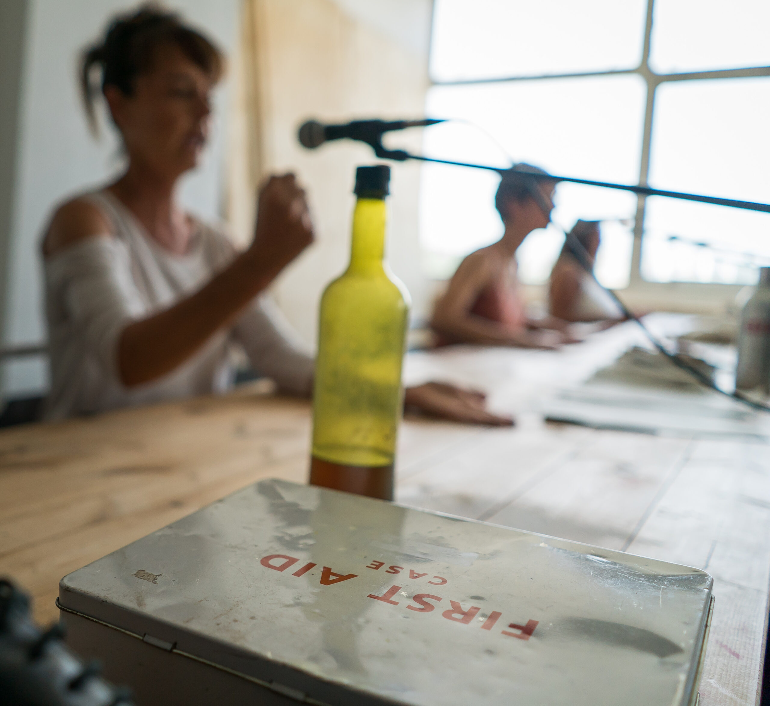 A person speaks into a microphone at a wooden table with a first aid case and bottle in focus. The setting is relaxed and casual.