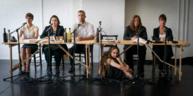 Six people sit at a wooden table with microphones and bottles, in a serious setting. A woman sits on the floor beneath, creating a sombre tone.