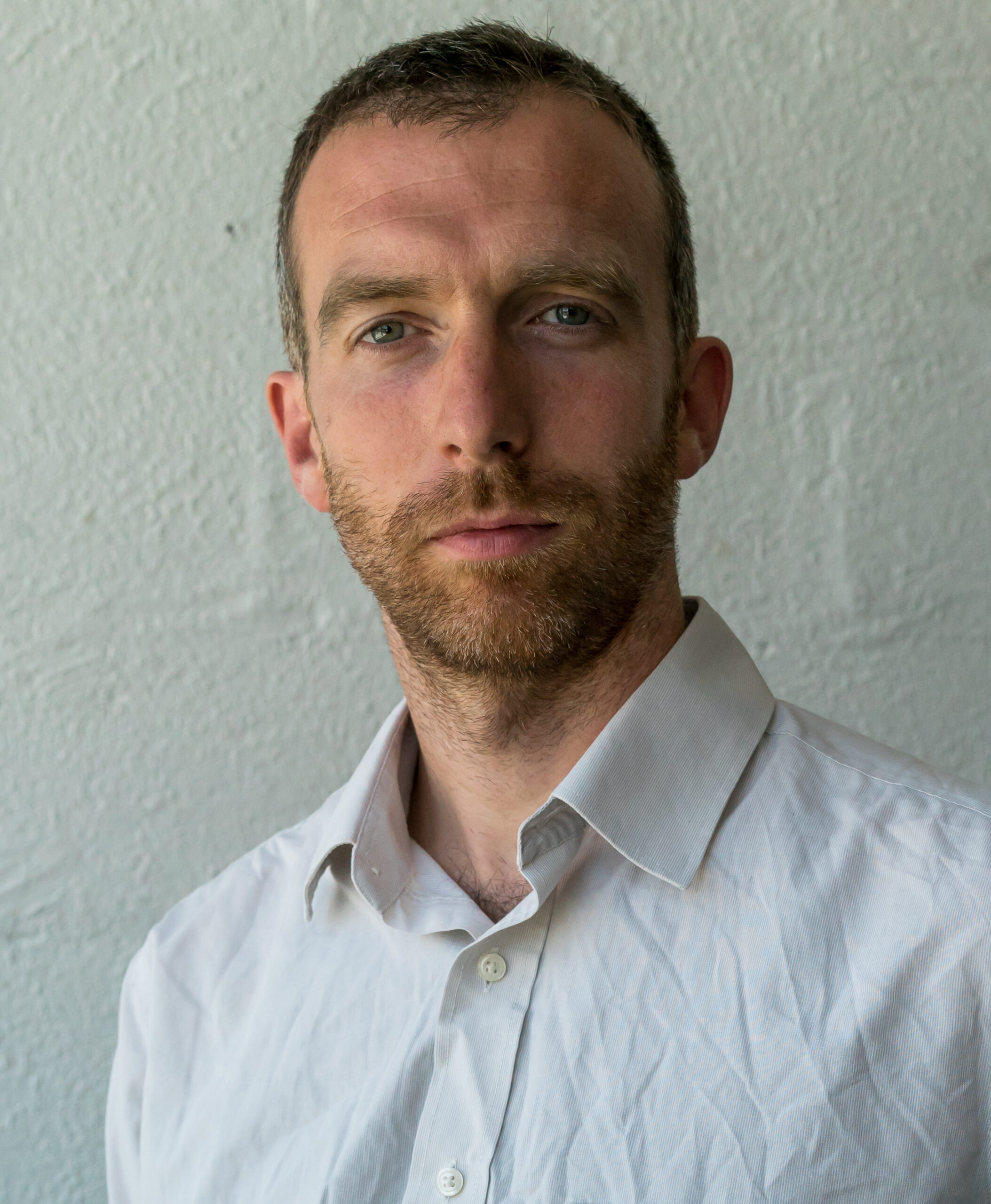 A man with short hair and beard looks at the camera with a neutral expression. He wears a white button-up shirt, standing against a plain light grey wall.