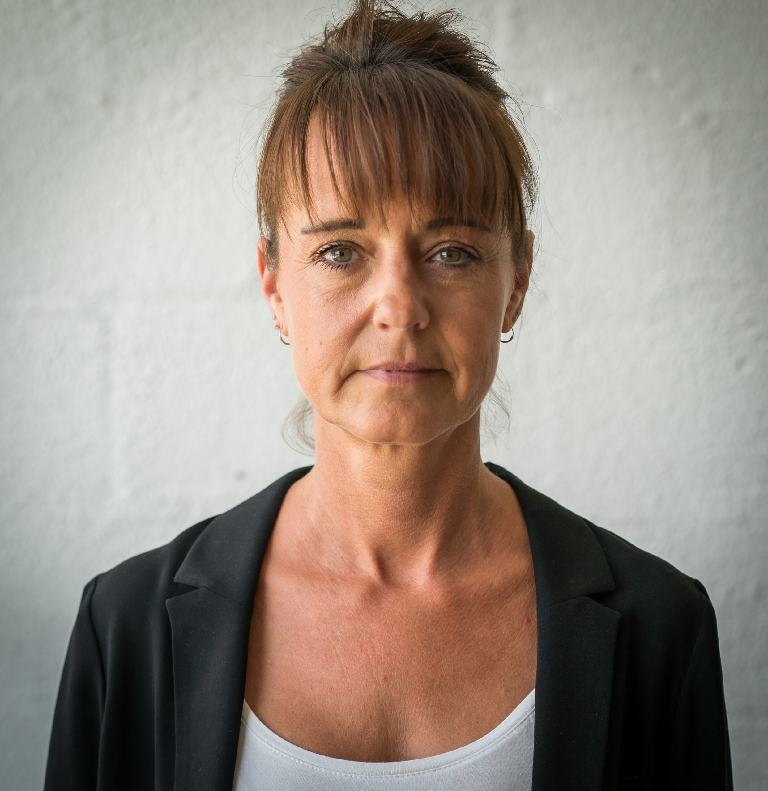 A woman with brown hair stands against a textured white wall, wearing a black blazer and white top. Her expression is calm and neutral.