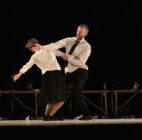 A man and woman perform a contemporary dance on stage, dressed in formal attire. The woman leans back as the man supports her, conveying tension and grace.