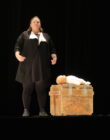 A performer dressed in black stands on stage beside a wooden crate, emotionless, with a doll wrapped in white lying on top. The scene is dramatic and intense.