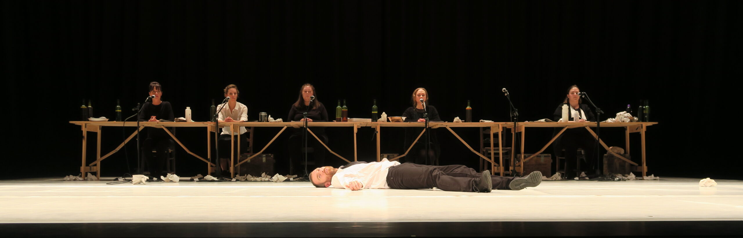 A person lies motionless on a stage, surrounded by six seated individuals at a long table with microphones, suggesting a dramatic performance scene.