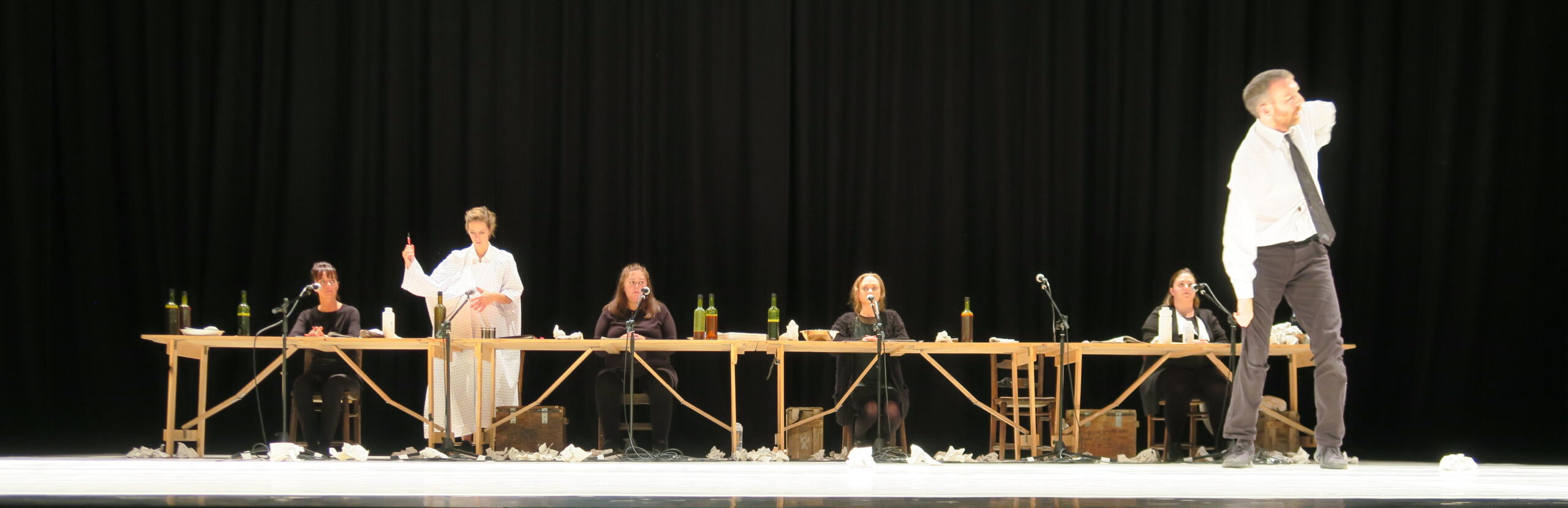 Six performers sit at a long table with microphones and bottles, while a seventh person stands off to the right in a theatrical pose. The stage is set with a dark curtain backdrop, and paper is scattered on the floor, conveying a dramatic and intense atmosphere.
