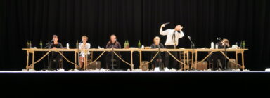 Seven people in dark attire sit at a long wooden table on stage, surrounded by wine bottles and papers, conveying a theatrical scene with dramatic gestures.