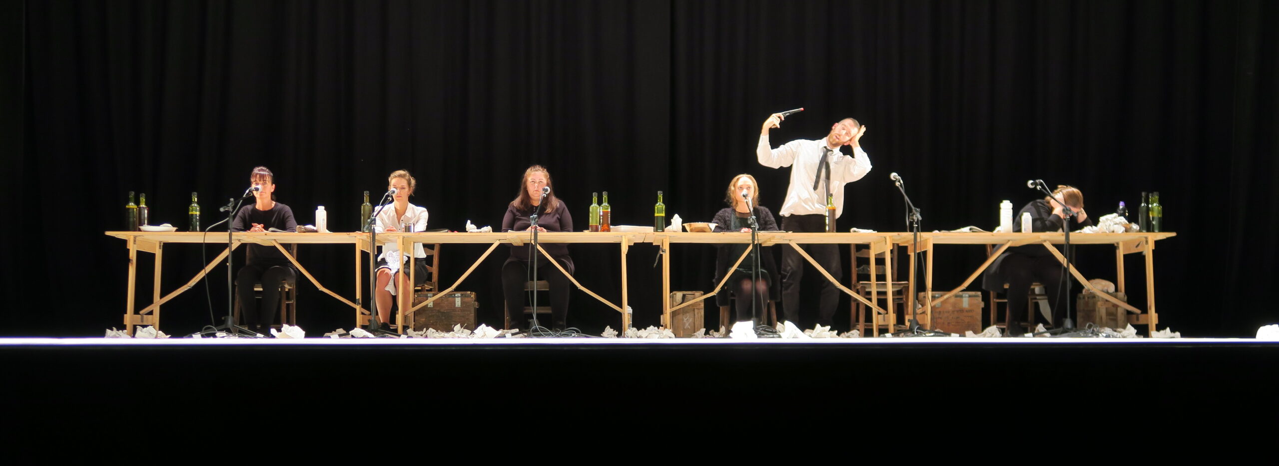 Seven people in dark attire sit at a long wooden table on stage, surrounded by wine bottles and papers, conveying a theatrical scene with dramatic gestures.