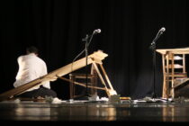 A man in a white shirt sits on a stage surrounded by broken wooden furniture and papers. Two microphones stand nearby, set against a dark backdrop.