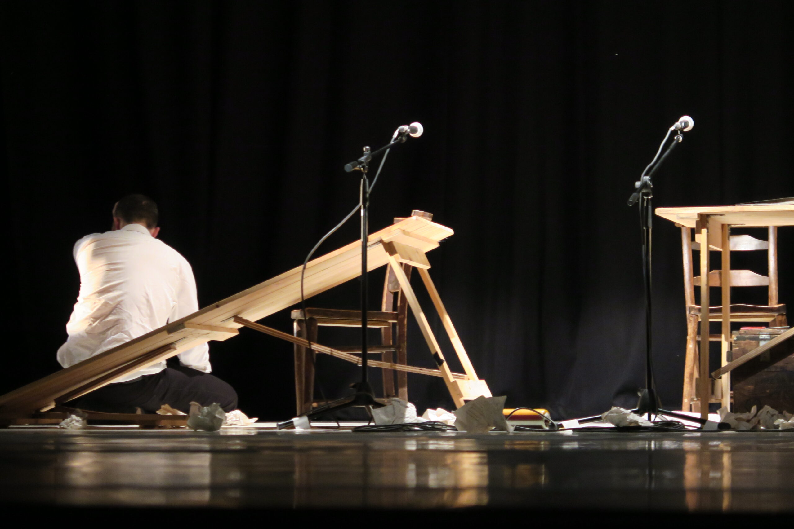 A man in a white shirt sits on a stage surrounded by broken wooden furniture and papers. Two microphones stand nearby, set against a dark backdrop.