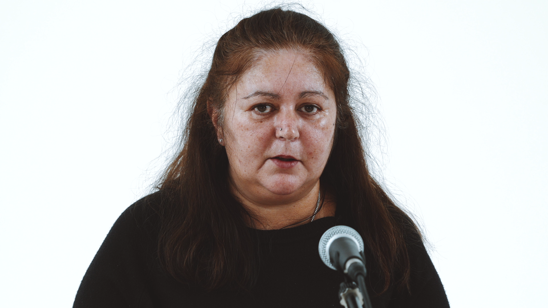 A woman with long dark hair speaks into a microphone, wearing a black top. The background is plain white, conveying a serious tone and focus.