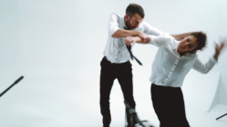 A man and woman in white shirts engage in a playful mock fight in a minimalistic studio setting, exuding a dynamic and energetic atmosphere.