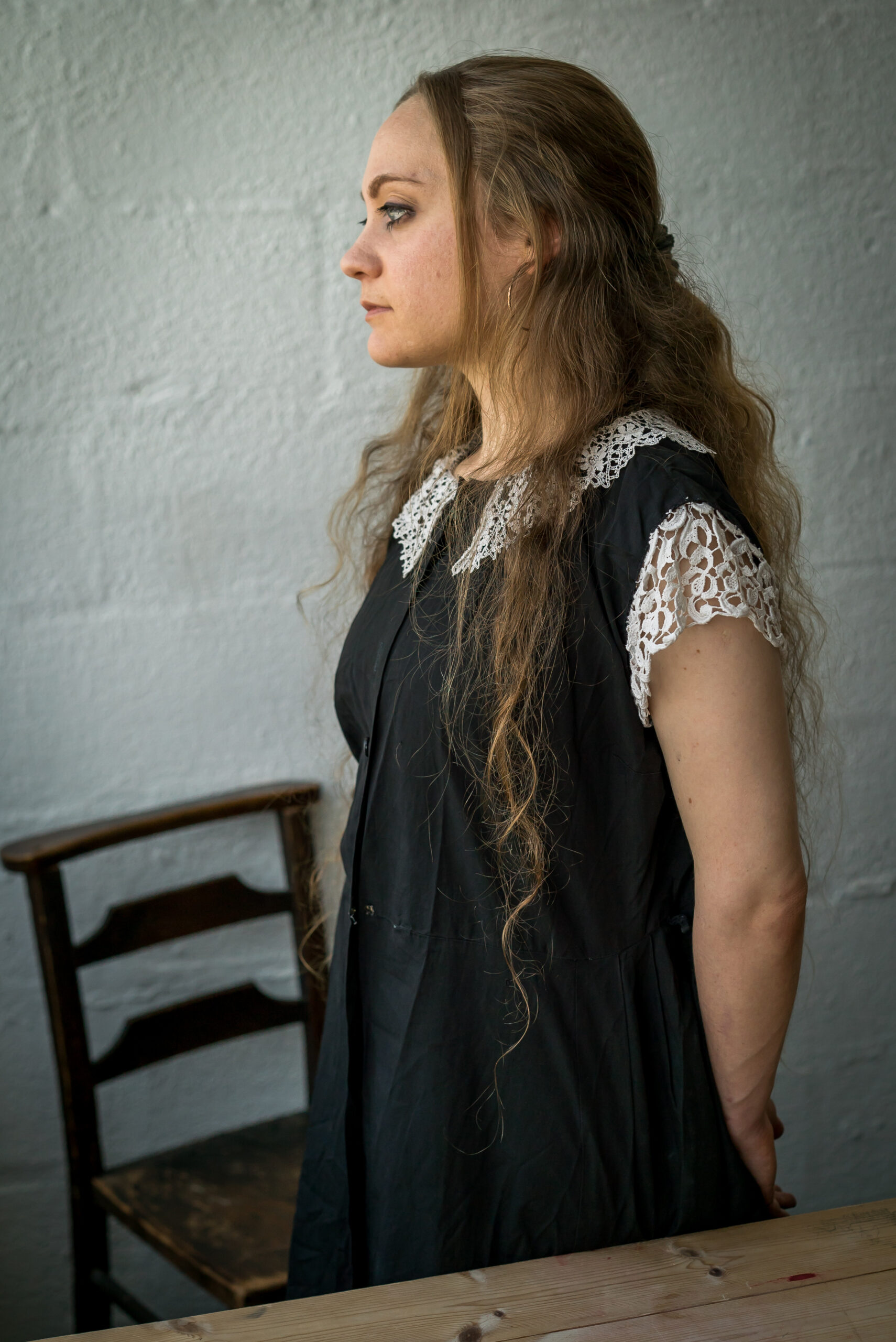 A woman in profile with long, wavy hair stands against a white wall. She's wearing a black dress with lace details, conveying a serene, vintage vibe. A wooden chair and table are partially visible.