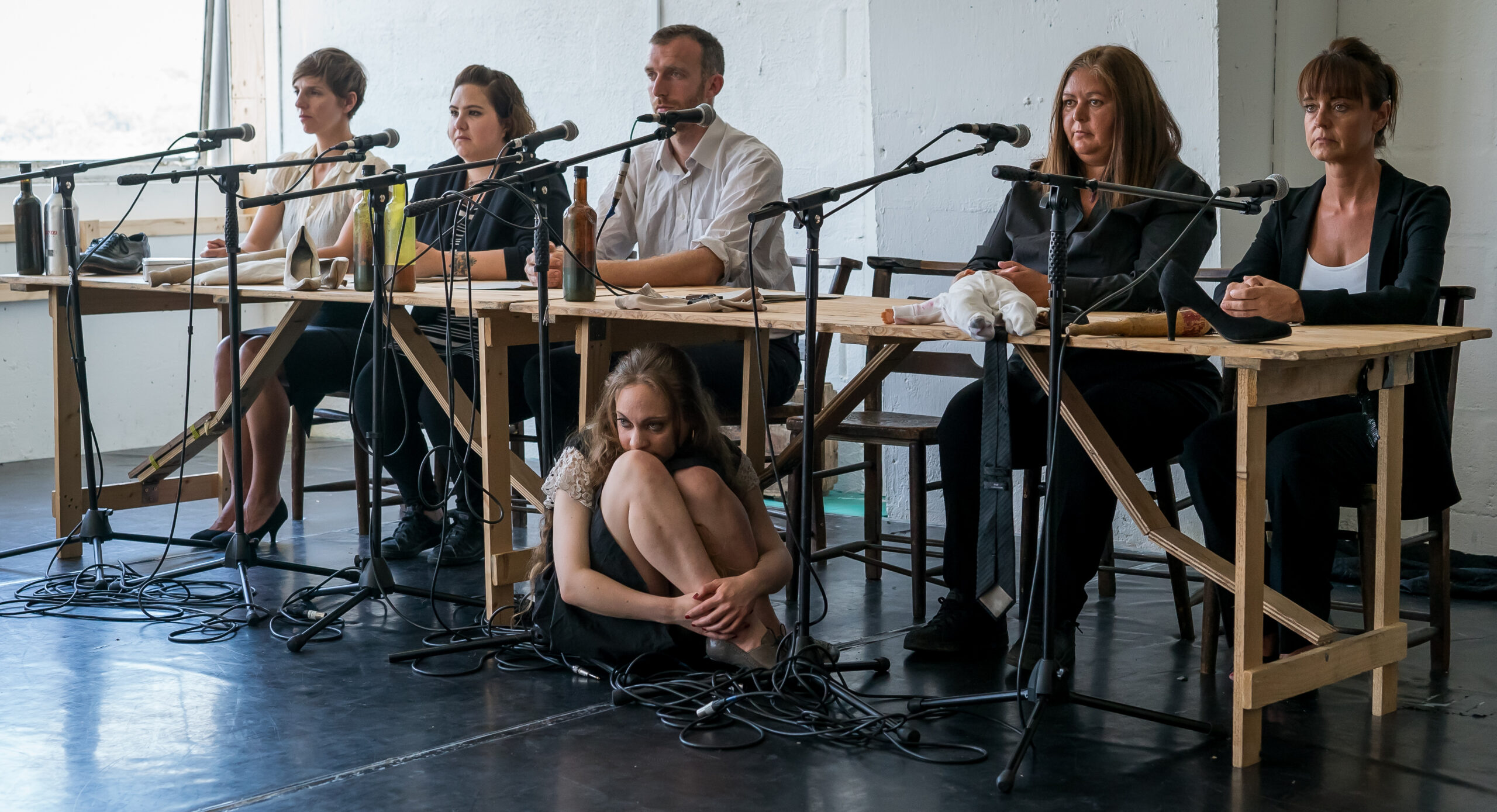 Six people seated at tables with microphones in front of them, staring intently. A woman sits on the floor, looking contemplative and reserved.