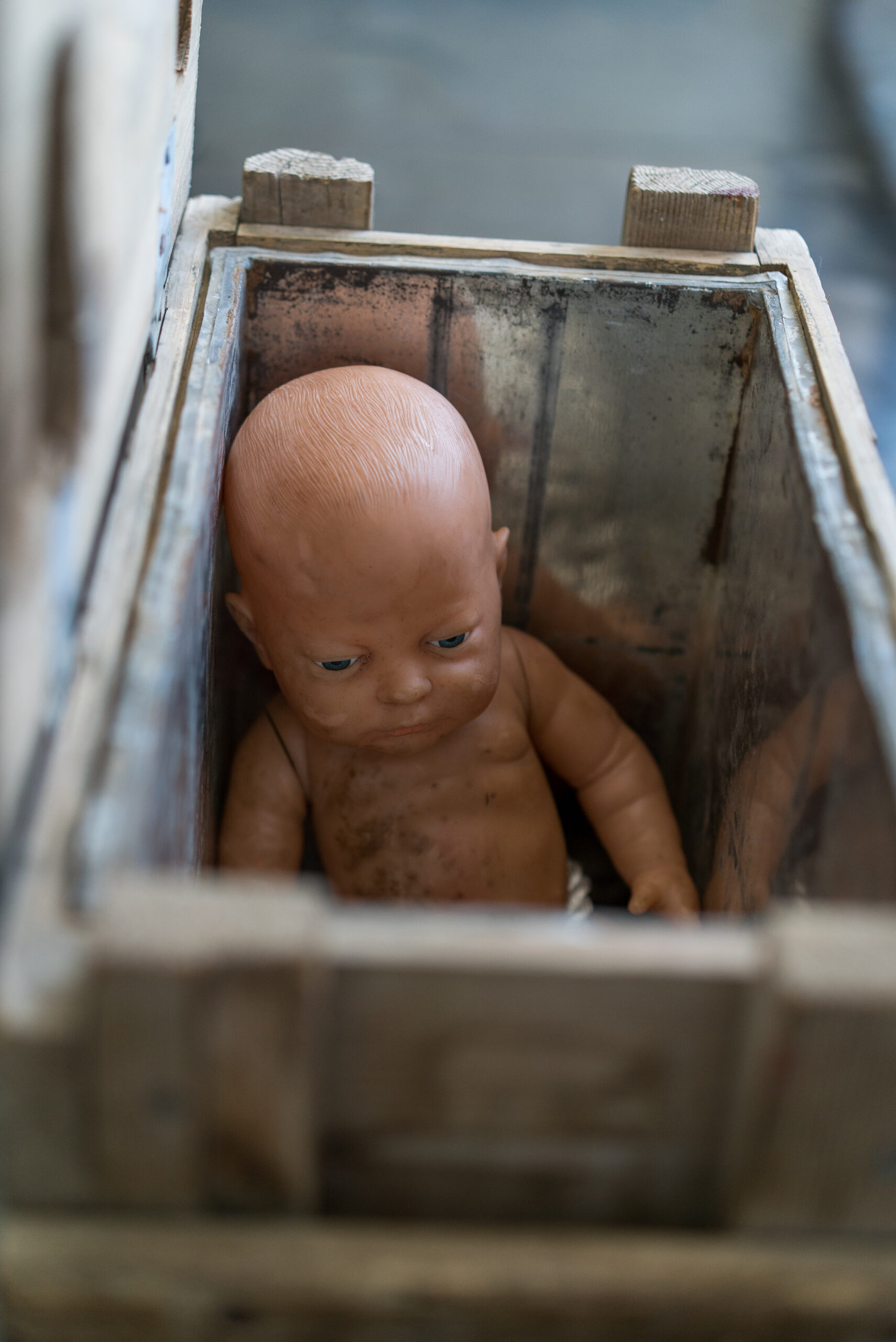 A worn-out baby doll with a bald head sits in an old, wooden box. The dim lighting and dusty setting convey an eerie, abandoned feel.