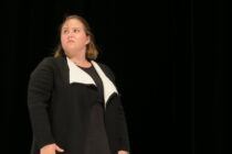 A person with short hair, wearing a black and white jacket, stands against a black background, looking slightly upward with a thoughtful expression.