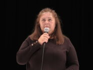 A woman stands on stage against a black background, holding a microphone. She appears focused and composed, wearing a long-sleeve dark top.