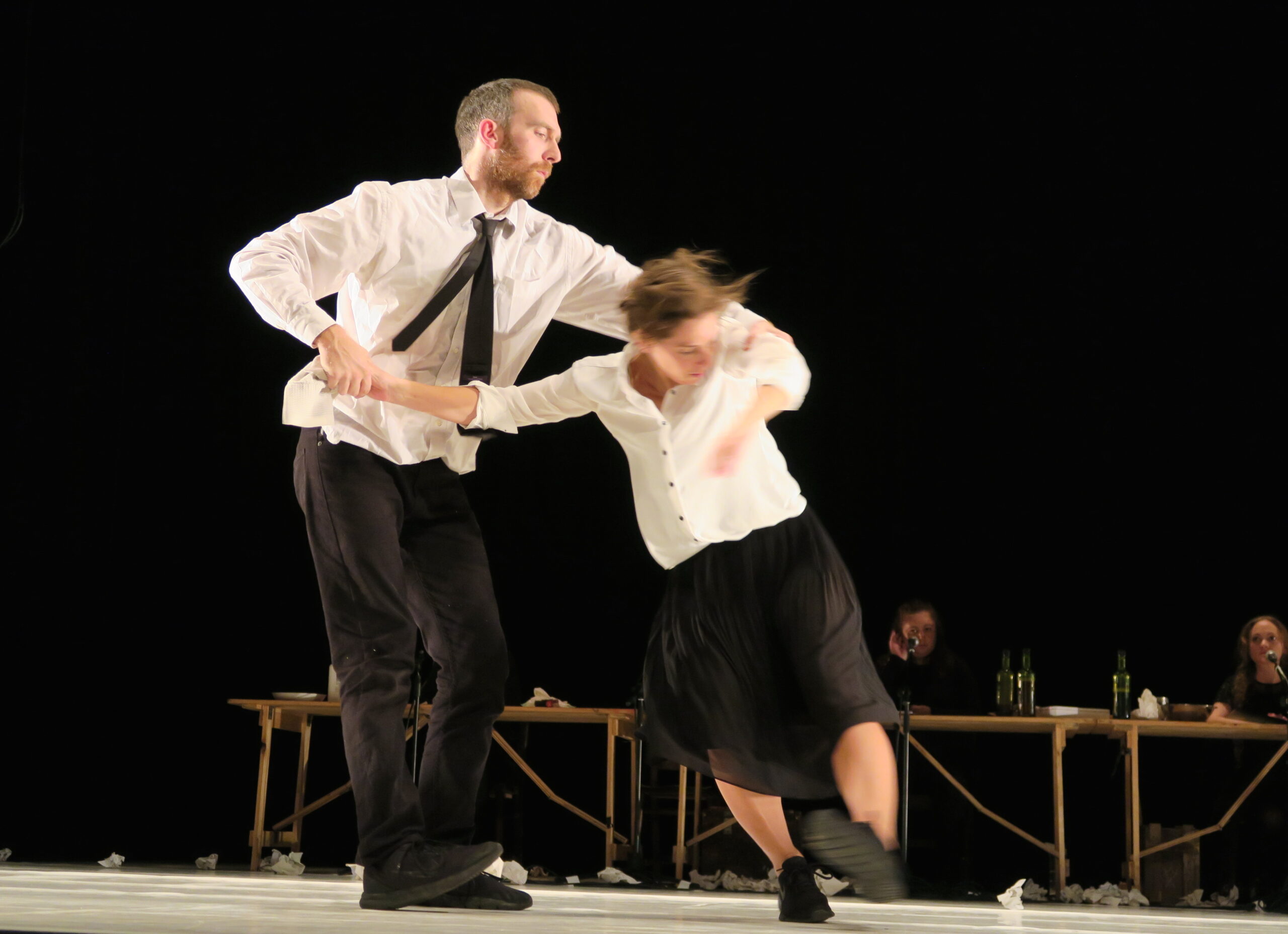 A man and a woman dance energetically on stage, both wearing white tops and black attire. Background features seated people at a table, set in a dark space.