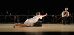 A woman in a white dress urgently reaches out toward a seated man on stage. Dimly lit figures and a long table form the backdrop, conveying tension.