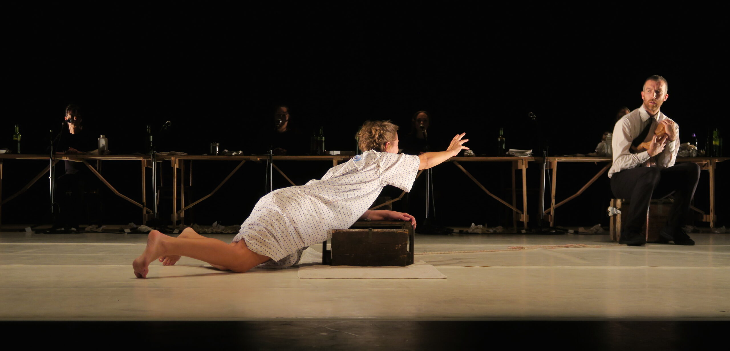 A woman in a white dress urgently reaches out toward a seated man on stage. Dimly lit figures and a long table form the backdrop, conveying tension.