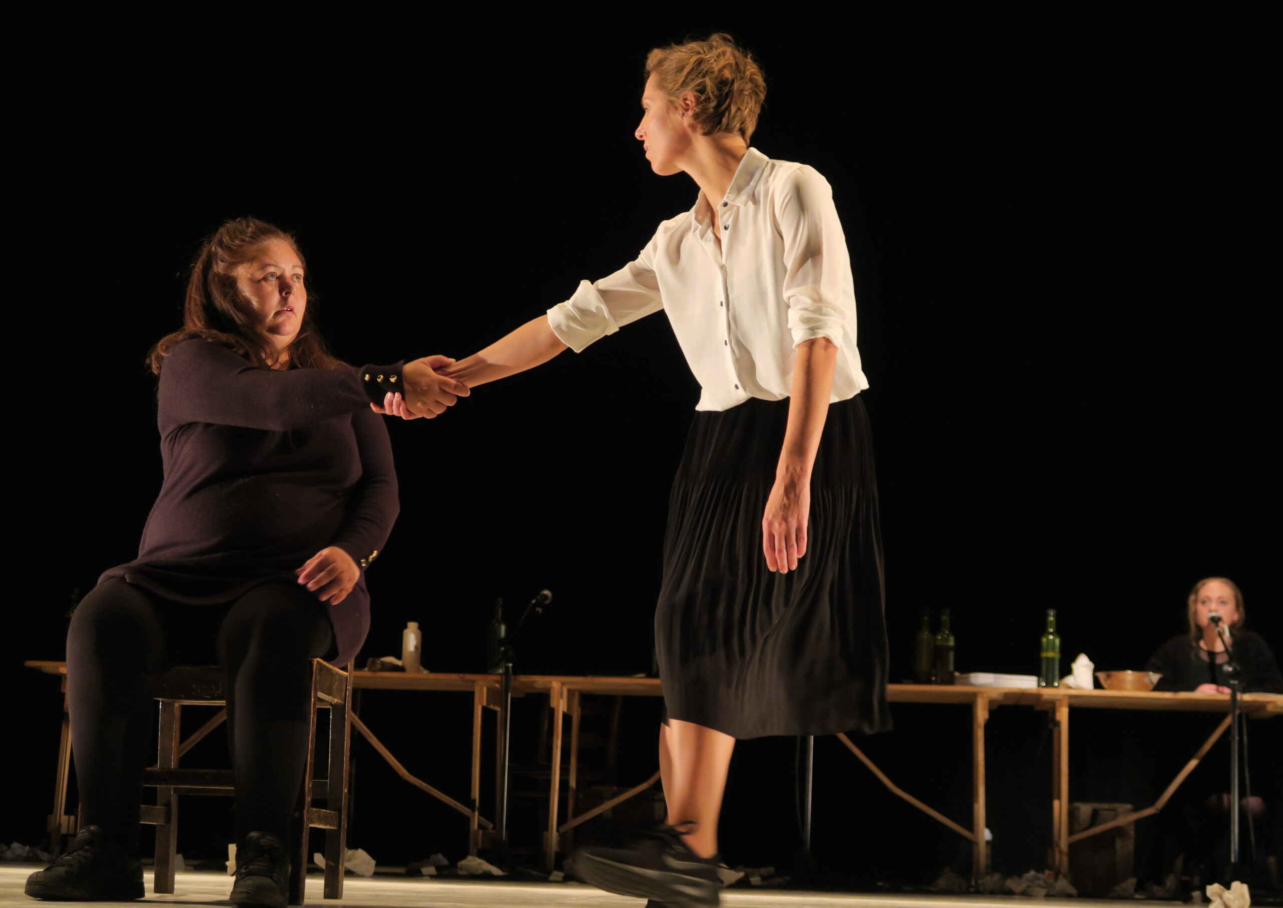 A woman sitting on a chair accepts a handshake from another standing woman in a white shirt and black skirt. A table with bottles is in the background.