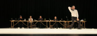 A man energetically dances in front of a long table where five seated individuals watch. The stage is decorated with bottles and papers, conveying a lively scene.