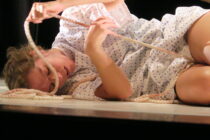 A person in a light patterned garment lies on the floor, loosely entangled in a rope, conveying a sense of vulnerability and introspection.