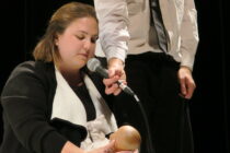 A woman kneels with her eyes closed, holding a microphone to a doll, assisted by a standing man. The scene is serious, with a focus on the interaction.
