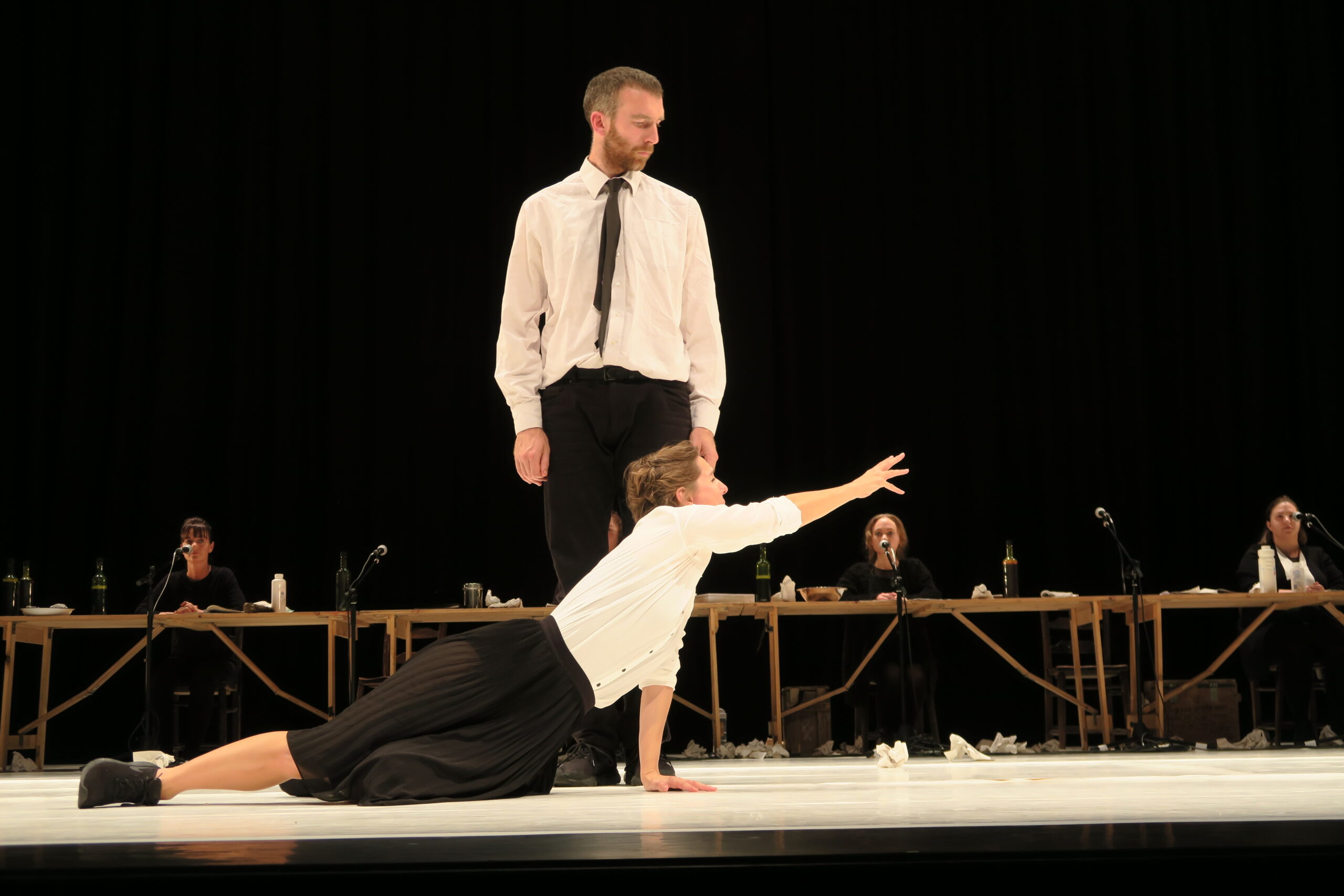 A person in a white shirt and black trousers stands while another in a matching outfit leans on one hand, reaching out. A table with people is in the background, creating a dramatic, intense atmosphere.