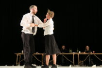 A man and woman in formal attire dance intensely onstage, conveying tension. A dimly lit table with people and bottles sits in the background.