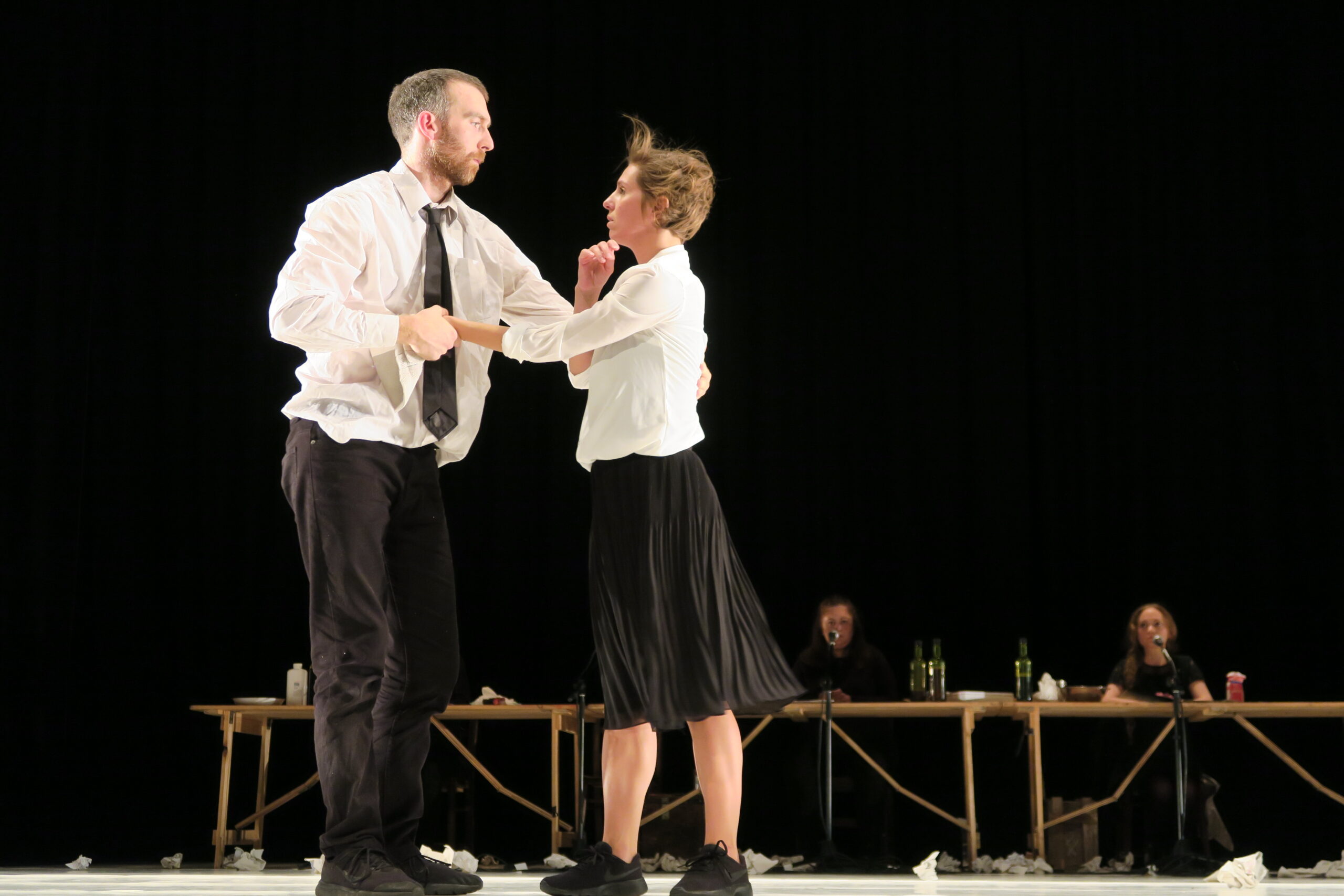 A man and woman in formal attire dance intensely onstage, conveying tension. A dimly lit table with people and bottles sits in the background.