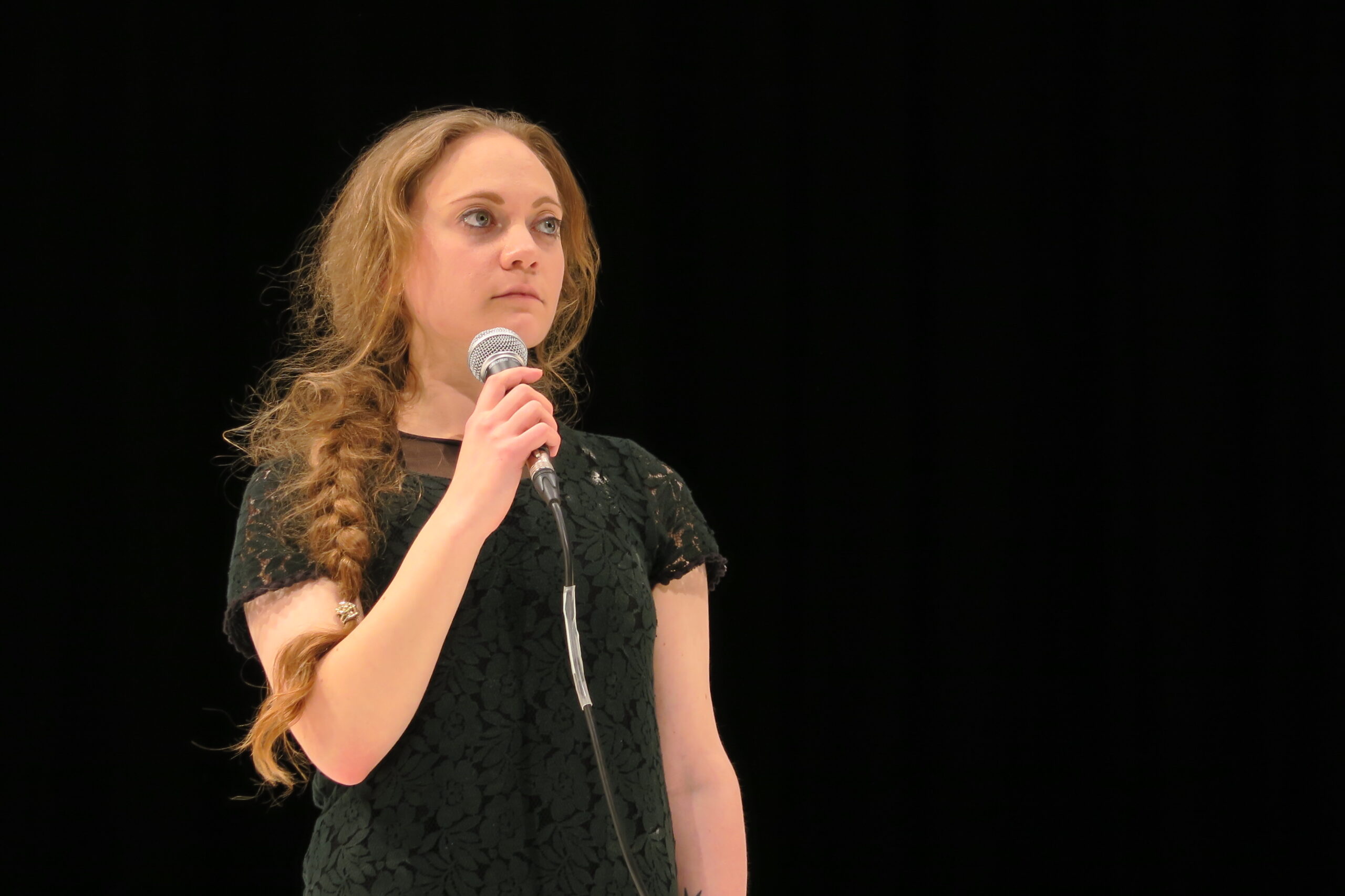 A woman with long braided hair stands against a black background, holding a microphone. Her expression is serious and focused, wearing a dark dress.