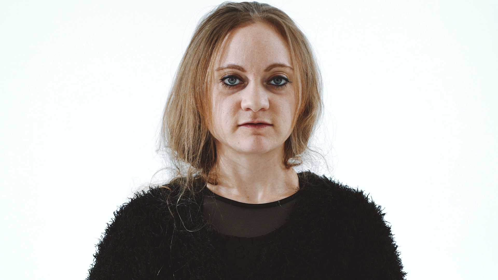 A woman with long, light brown hair, wearing a black fuzzy sweater, stands against a plain white background, looking directly ahead with a neutral expression.