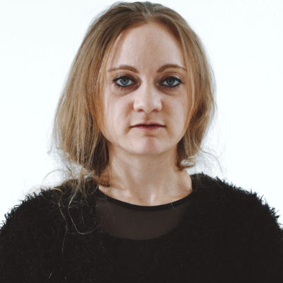 A woman with long hair and a neutral expression stands against a white background. She wears a black, textured sweater, conveying a calm, serene mood.