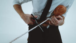 A person in a white shirt and black tie holds a damaged doll while cutting a rope with scissors. The scene conveys tension and urgency.